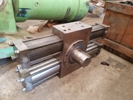 A large, industrial cylinder with multiple metallic rods and bolts is positioned on a concrete floor. The machinery appears robust and heavy-duty, likely used for mechanical or hydraulic purposes. Green and wooden structures are in the background, suggesting a workshop or manufacturing setting.