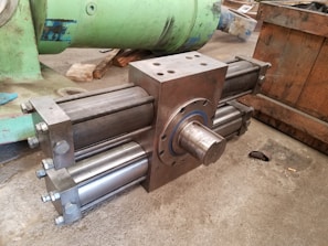 A large, industrial cylinder with multiple metallic rods and bolts is positioned on a concrete floor. The machinery appears robust and heavy-duty, likely used for mechanical or hydraulic purposes. Green and wooden structures are in the background, suggesting a workshop or manufacturing setting.