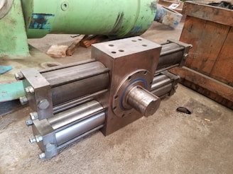 A large, industrial cylinder with multiple metallic rods and bolts is positioned on a concrete floor. The machinery appears robust and heavy-duty, likely used for mechanical or hydraulic purposes. Green and wooden structures are in the background, suggesting a workshop or manufacturing setting.