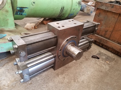 A large, industrial cylinder with multiple metallic rods and bolts is positioned on a concrete floor. The machinery appears robust and heavy-duty, likely used for mechanical or hydraulic purposes. Green and wooden structures are in the background, suggesting a workshop or manufacturing setting.