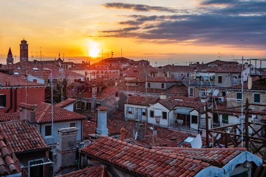 A picturesque view of a city with red-tiled rooftops, silhouetted buildings, and a towering bell tower under a vibrant sky at sunset. The sun is setting on the horizon, casting warm orange and gold hues across the sky and reflecting on the rooftops. Some antennas and laundry drying on a rooftop are visible, enhancing the urban landscape.
