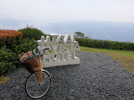 A scenic viewpoint overlooks a body of water and distant mountains, featuring a large white sign with the text '#TAAL VISTA HOTEL'. In the foreground, a vintage-style bicycle with a basket of colorful flowers is parked on a gravel path. Surrounding the area are neatly manicured bushes and grass.