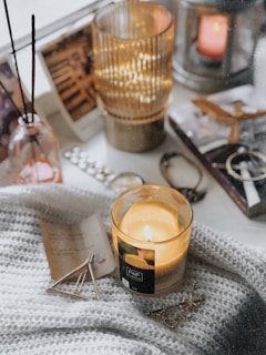 A cozy setting featuring a lit candle in a glass container, surrounded by various items such as a knitted blanket, a small pile of white sticks or reeds in a glass bottle, books, a photograph, an ornamental lantern holding another candle, some jewelry including bracelets and a chain, and a small folded piece of paper.