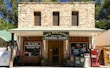 A rustic stone building houses a general store named M & M Mercantile. The storefront features a green metal roof and a large sign advertising wine, spirits, ice cream, and various other goods and services. Two large windows on the upper floor add to the building's old-fashioned aesthetic. An open sign and various vending machines, including one for Coca-Cola, are present near the entrance.