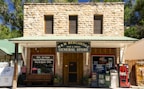 A rustic stone building houses a general store named M & M Mercantile. The storefront features a green metal roof and a large sign advertising wine, spirits, ice cream, and various other goods and services. Two large windows on the upper floor add to the building's old-fashioned aesthetic. An open sign and various vending machines, including one for Coca-Cola, are present near the entrance.