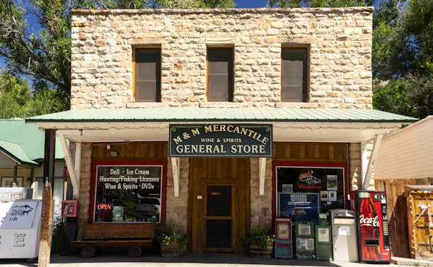 A rustic stone building houses a general store named M & M Mercantile. The storefront features a green metal roof and a large sign advertising wine, spirits, ice cream, and various other goods and services. Two large windows on the upper floor add to the building's old-fashioned aesthetic. An open sign and various vending machines, including one for Coca-Cola, are present near the entrance.