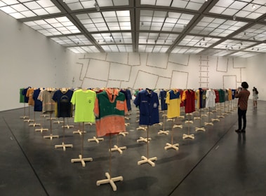 A large room with a grid-like ceiling and grey flooring displays an installation of various sports jerseys hung on wooden stands arranged in rows. People are observing and photographing the exhibit.