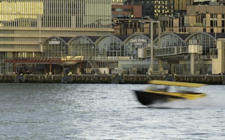 A waterfront scene featuring a modern building labeled 'Cruise Terminal Rotterdam' with large arched windows. The foreground shows a yellow and black boat speeding across the water, creating a motion blur effect. The building's architecture includes multiple levels and a glass facade. The water appears calm despite the movement of the boat.