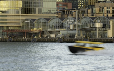 A waterfront scene featuring a modern building labeled 'Cruise Terminal Rotterdam' with large arched windows. The foreground shows a yellow and black boat speeding across the water, creating a motion blur effect. The building's architecture includes multiple levels and a glass facade. The water appears calm despite the movement of the boat.