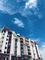 A multi-story residential building featuring a mix of red, beige, and white colors along with large white columns. The sky in the background is bright and blue with scattered white clouds. Some utility wires are visible in the foreground.