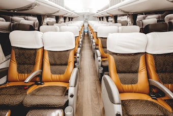 An empty interior of a modern bus or coach with rows of upholstered seats featuring white headrest covers and patterned cushions. The aisle is clear, and the interior appears clean and well-maintained, with overhead compartments visible and muted lighting.