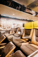 The interior of a modern bus with spacious, reclining seats covered in patterned upholstery. The seats have individual armrests and controls, and the overhead area is well-lit with spotlights. The curtains along the windows create a cozy atmosphere.