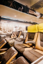 The interior of a modern bus with spacious, reclining seats covered in patterned upholstery. The seats have individual armrests and controls, and the overhead area is well-lit with spotlights. The curtains along the windows create a cozy atmosphere.
