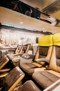The interior of a modern bus with spacious, reclining seats covered in patterned upholstery. The seats have individual armrests and controls, and the overhead area is well-lit with spotlights. The curtains along the windows create a cozy atmosphere.