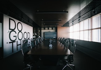 A modern conference room with a long wooden table surrounded by mesh office chairs. On the left wall, large bold letters spell out 'Do Good Things'. The room is well-lit by natural light coming through Venetian blinds on the right side. Overhead, there are ceiling lights and a grid-like ceiling design. The room has a minimalist and professional decor.