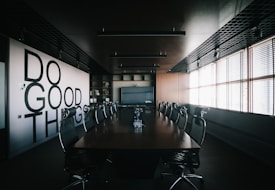 A modern conference room with a long wooden table surrounded by mesh office chairs. On the left wall, large bold letters spell out 'Do Good Things'. The room is well-lit by natural light coming through Venetian blinds on the right side. Overhead, there are ceiling lights and a grid-like ceiling design. The room has a minimalist and professional decor.