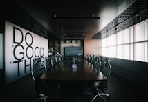 A modern conference room with a long wooden table surrounded by mesh office chairs. On the left wall, large bold letters spell out 'Do Good Things'. The room is well-lit by natural light coming through Venetian blinds on the right side. Overhead, there are ceiling lights and a grid-like ceiling design. The room has a minimalist and professional decor.