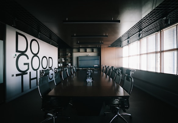 A modern conference room with a long wooden table surrounded by mesh office chairs. On the left wall, large bold letters spell out 'Do Good Things'. The room is well-lit by natural light coming through Venetian blinds on the right side. Overhead, there are ceiling lights and a grid-like ceiling design. The room has a minimalist and professional decor.