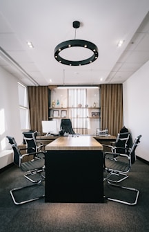 A modern office with a long wooden conference table surrounded by black chairs. The back wall features wooden slats and shelves displaying trophies and decorative items. A circular ceiling light hangs above the table, and natural light enters from windows on the side.