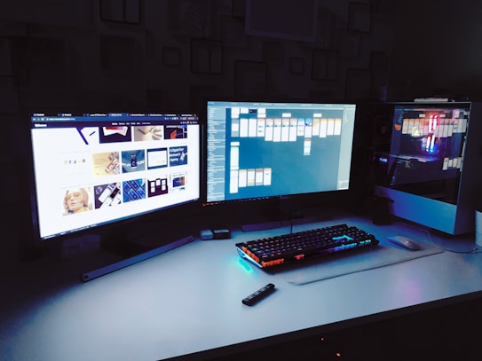 A modern desktop setup with three monitors displaying different content, one showing a website and another displaying a flowchart or workflow application. The ambient lighting from the keyboard and the computer&rsquo;s internals adds a colorful glow to the dark room.