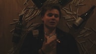 A person lies on a wooden surface surrounded by multiple cigarettes and three wine bottles. The individual holds a lit cigarette and wears a cross necklace.