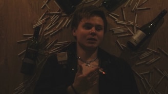 A person lies on a wooden surface surrounded by multiple cigarettes and three wine bottles. The individual holds a lit cigarette and wears a cross necklace.