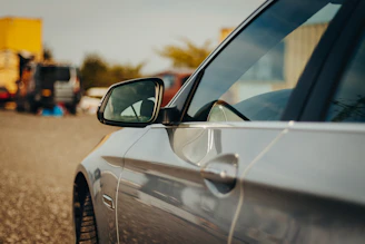 A close-up view of a sleek car door and side mirror with a slight reflection. The background shows a blurred scene with vehicles and containers, suggesting a parking lot or industrial area.