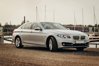 A silver luxury sedan is parked on a gravel surface near a dock. The background includes several boats and a building, suggesting a waterside location. The vehicle's design is sleek and modern with visible front grilles and headlights.