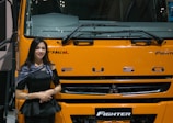 A woman stands beside the front of a large yellow-orange vehicle, displaying the brand name 'FUSO' prominently. She is wearing a black outfit and a shawl with patterns, smiling slightly. The vehicle is labeled 'Fighter' and is likely part of a display or exhibition setting.