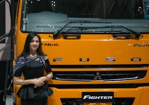 A woman stands beside the front of a large yellow-orange vehicle, displaying the brand name 'FUSO' prominently. She is wearing a black outfit and a shawl with patterns, smiling slightly. The vehicle is labeled 'Fighter' and is likely part of a display or exhibition setting.