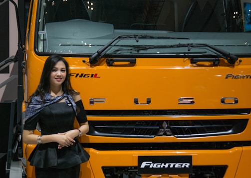 A woman stands beside the front of a large yellow-orange vehicle, displaying the brand name 'FUSO' prominently. She is wearing a black outfit and a shawl with patterns, smiling slightly. The vehicle is labeled 'Fighter' and is likely part of a display or exhibition setting.