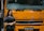 A woman stands beside the front of a large yellow-orange vehicle, displaying the brand name 'FUSO' prominently. She is wearing a black outfit and a shawl with patterns, smiling slightly. The vehicle is labeled 'Fighter' and is likely part of a display or exhibition setting.