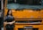 A woman stands beside the front of a large yellow-orange vehicle, displaying the brand name 'FUSO' prominently. She is wearing a black outfit and a shawl with patterns, smiling slightly. The vehicle is labeled 'Fighter' and is likely part of a display or exhibition setting.