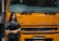 A woman stands beside the front of a large yellow-orange vehicle, displaying the brand name 'FUSO' prominently. She is wearing a black outfit and a shawl with patterns, smiling slightly. The vehicle is labeled 'Fighter' and is likely part of a display or exhibition setting.