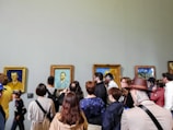 A group of people is gathered in an art gallery observing several framed paintings on the wall. The paintings include portraits and landscapes with wooden frames. The attendees are of varying ages, some taking photos, others discussing or simply looking. The scene captures a diverse mix of casual and formally dressed visitors.