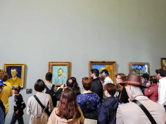 A group of people is gathered in an art gallery observing several framed paintings on the wall. The paintings include portraits and landscapes with wooden frames. The attendees are of varying ages, some taking photos, others discussing or simply looking. The scene captures a diverse mix of casual and formally dressed visitors.