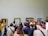 A group of people is gathered in an art gallery observing several framed paintings on the wall. The paintings include portraits and landscapes with wooden frames. The attendees are of varying ages, some taking photos, others discussing or simply looking. The scene captures a diverse mix of casual and formally dressed visitors.