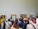 A group of people is gathered in an art gallery observing several framed paintings on the wall. The paintings include portraits and landscapes with wooden frames. The attendees are of varying ages, some taking photos, others discussing or simply looking. The scene captures a diverse mix of casual and formally dressed visitors.