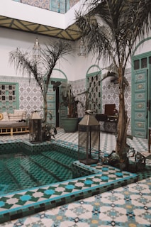 A beautifully designed indoor courtyard featuring intricately patterned tiles in shades of teal, blue, and white. Tall potted plants add a touch of greenery next to a small pool with steps leading into it. The surrounding walls display detailed tile work and ornate doors with a similar color scheme. Lantern-style lights hang from the ceiling, enhancing the atmosphere of serenity and elegance.