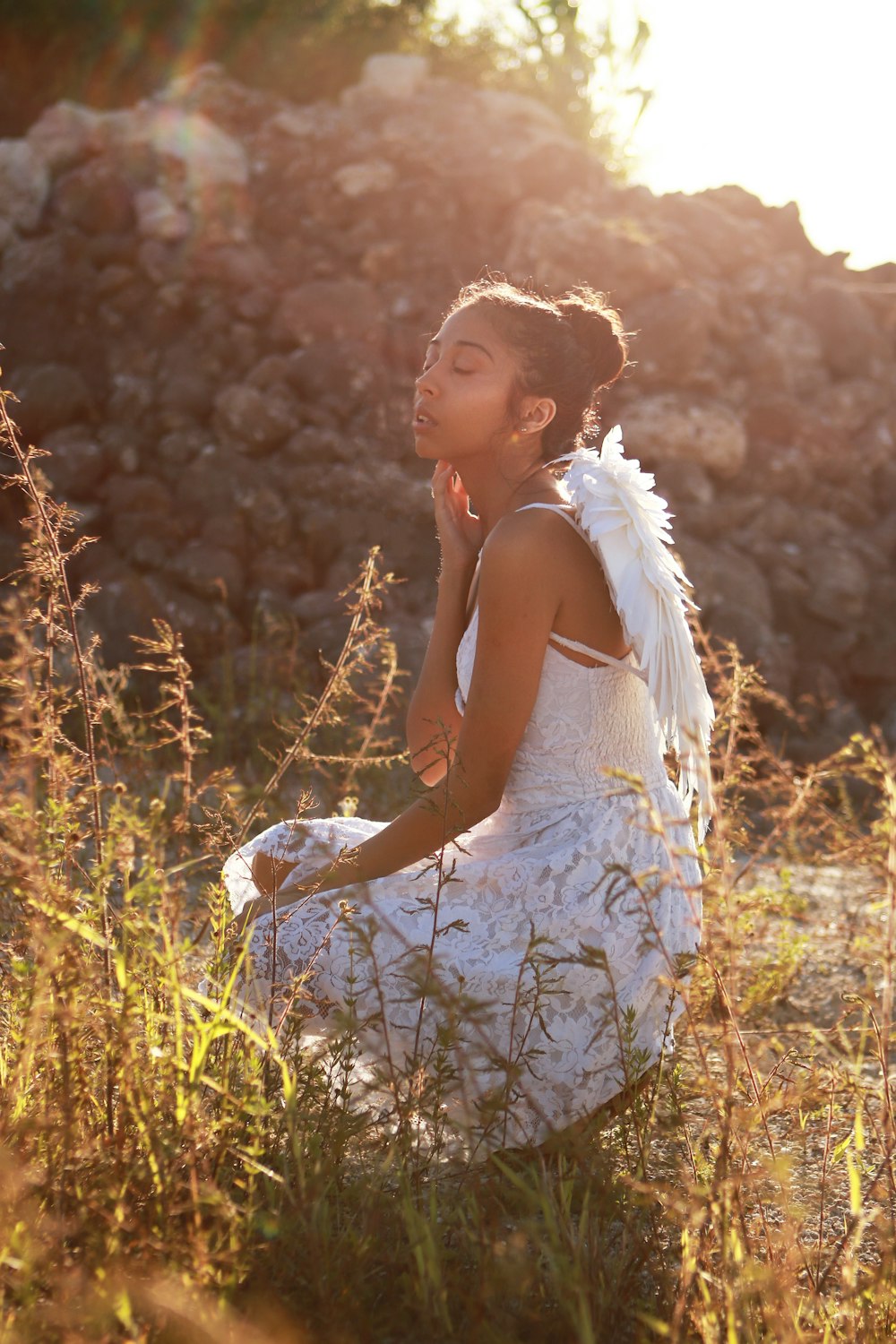 woman wearing white spaghetti strap dress with wings touching her neck while sitting near green field during daytime