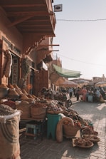 A bustling outdoor market scene with baskets filled with spices and goods lining the cobblestone pathway. The market stall is covered by a wooden awning and shoppers are visible in the background, some sitting at small tables. Umbrellas provide shade from the sunlight, casting soft shadows.
