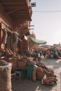 A bustling outdoor market scene with baskets filled with spices and goods lining the cobblestone pathway. The market stall is covered by a wooden awning and shoppers are visible in the background, some sitting at small tables. Umbrellas provide shade from the sunlight, casting soft shadows.