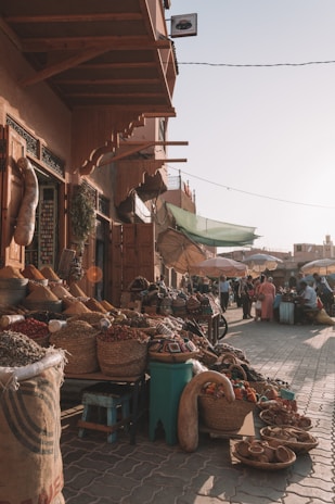 A bustling outdoor market scene with baskets filled with spices and goods lining the cobblestone pathway. The market stall is covered by a wooden awning and shoppers are visible in the background, some sitting at small tables. Umbrellas provide shade from the sunlight, casting soft shadows.