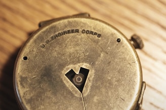 An old, worn metallic device featuring the words 'U.S. ENGINEER CORPS' engraved on it. The device has visible scratches and a circular shape with a small winding knob on the side. The surface has a slightly tarnished appearance, set against a wooden background.