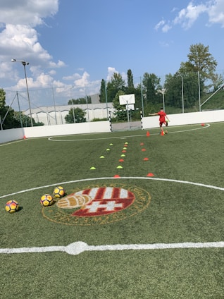 A person in sports attire is on a synthetic grass football field, practicing with cones laid out for drills. Three footballs are placed on the ground. A logo is visible in the center circle of the field, and a basketball hoop stands attached to the goal post. Trees and a partially cloudy sky are in the background.