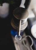 A close-up of a metal dispenser with a sleek, minimalist design. The image is focused on the pump handle, with a clear drop pending to fall from it. There is a blurred background with subtle hints of other objects and light sources.