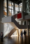 A modern architectural interior with large, angular windows allowing natural light to flood in. A central, curved staircase leads to an upper level, where two people stand looking out over the space. Below, three people are walking and interacting in the open area. The design features clean lines and a prominent red square artwork on the wall.