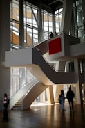 A modern architectural interior with large, angular windows allowing natural light to flood in. A central, curved staircase leads to an upper level, where two people stand looking out over the space. Below, three people are walking and interacting in the open area. The design features clean lines and a prominent red square artwork on the wall.