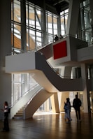 A modern architectural interior with large, angular windows allowing natural light to flood in. A central, curved staircase leads to an upper level, where two people stand looking out over the space. Below, three people are walking and interacting in the open area. The design features clean lines and a prominent red square artwork on the wall.