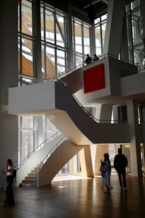 A modern architectural interior with large, angular windows allowing natural light to flood in. A central, curved staircase leads to an upper level, where two people stand looking out over the space. Below, three people are walking and interacting in the open area. The design features clean lines and a prominent red square artwork on the wall.